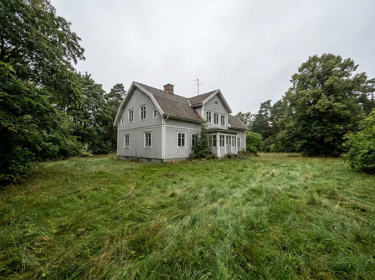 Grått trähus omgivet av grönska och lång unkempt gräsmatta.
